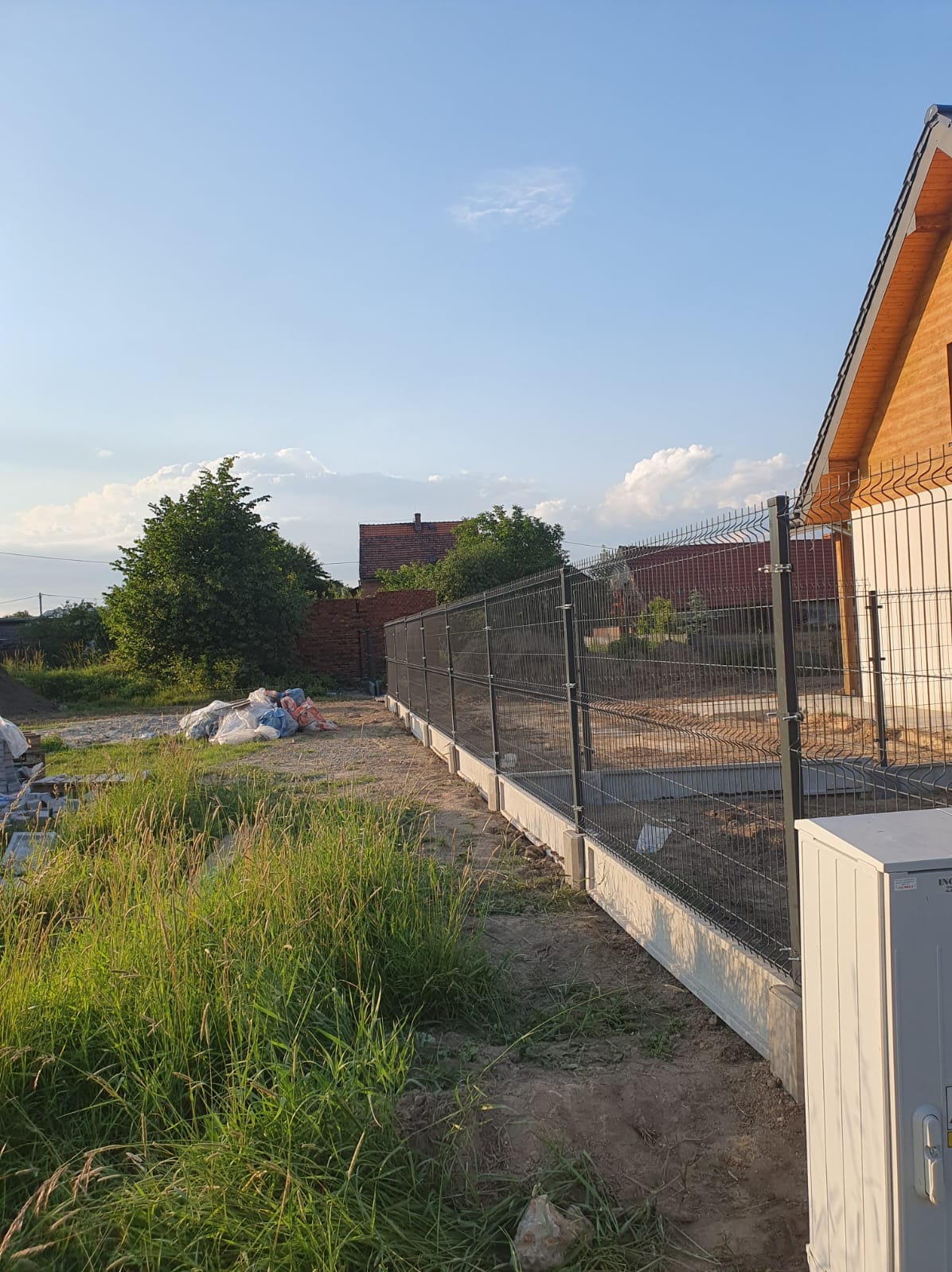 Nowo postawione ogrodzenie panelowe z podmurówką betonową, widoczne worki z materiałami budowlanymi w tle, wysoka trawa z przodu, dom z cegły i drewniany dom w oddali.