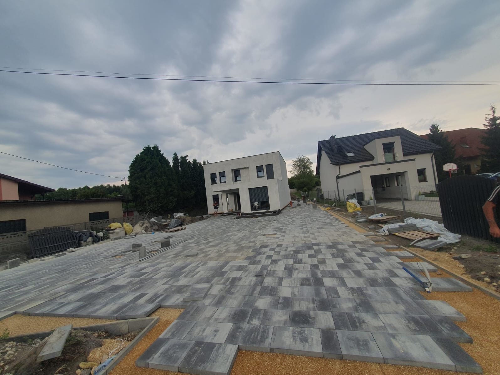 Fragment podjazdu z szarej kostki brukowej ułożonej we wzór jodełki, widoczne dwa nowoczesne domy w tle, etap prac w trakcie układania, podsypka piaskowa.