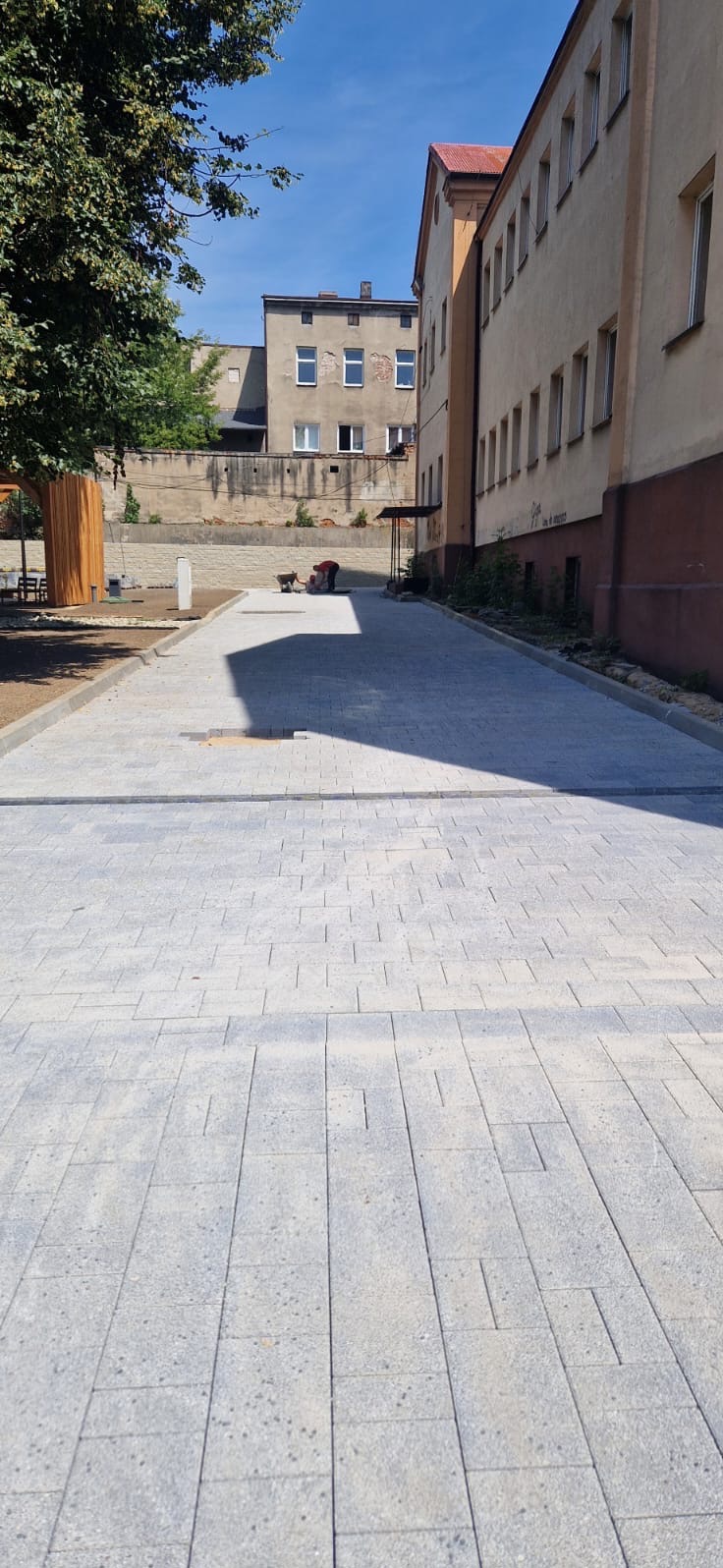 Świeżo ułożona, szara kostka brukowa na placu budowy w słoneczny dzień, z pracownikiem i budynkami w tle.