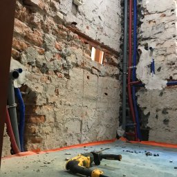 WERMAXPOL - Remont łazienki w trakcie: surowe ściany z cegły i betonu, widoczne rury instalacji wodnej i elektrycznej, wiertarka DeWalt leżąca na podłodze z folią ochronną.