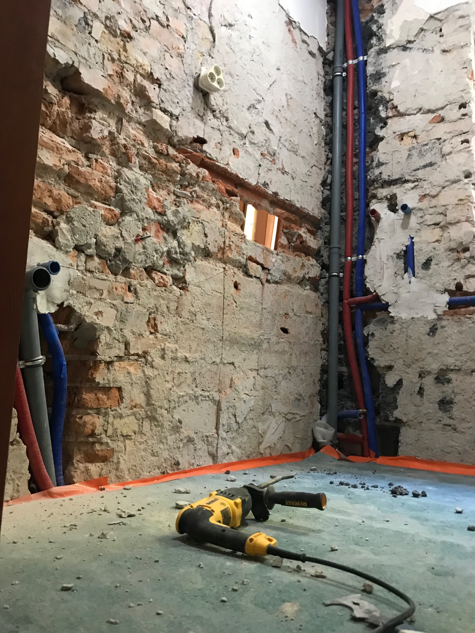 Remont łazienki w trakcie: surowe ściany z cegły i betonu, widoczne rury instalacji wodnej i elektrycznej, wiertarka DeWalt leżąca na podłodze z folią ochronną.