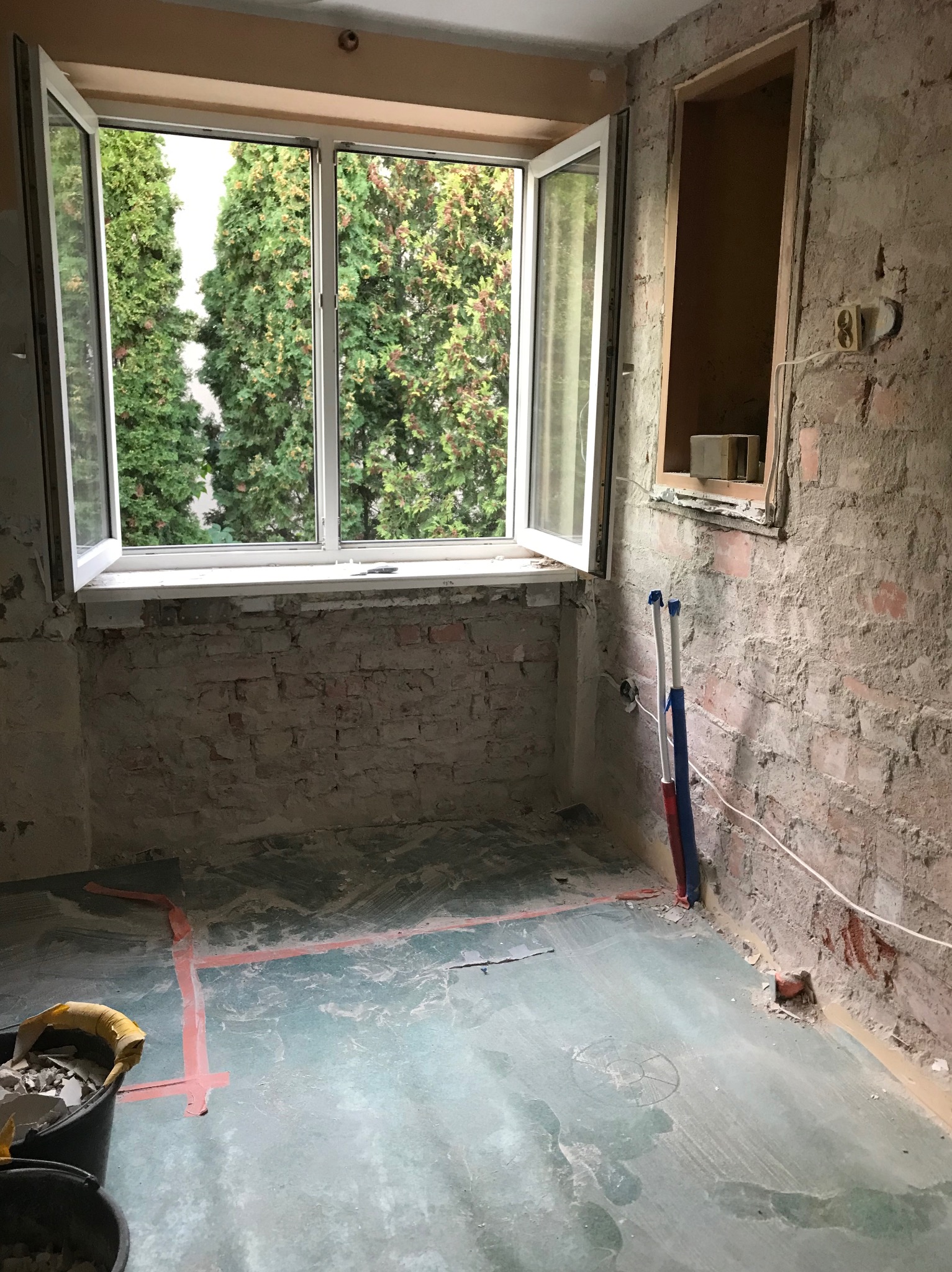 Pokój w trakcie remontu: odsłonięta cegła na ścianach, otwarte okno z widokiem na zieleń, widoczne rury instalacyjne i ślady po wyznaczonych liniach na podłodze.