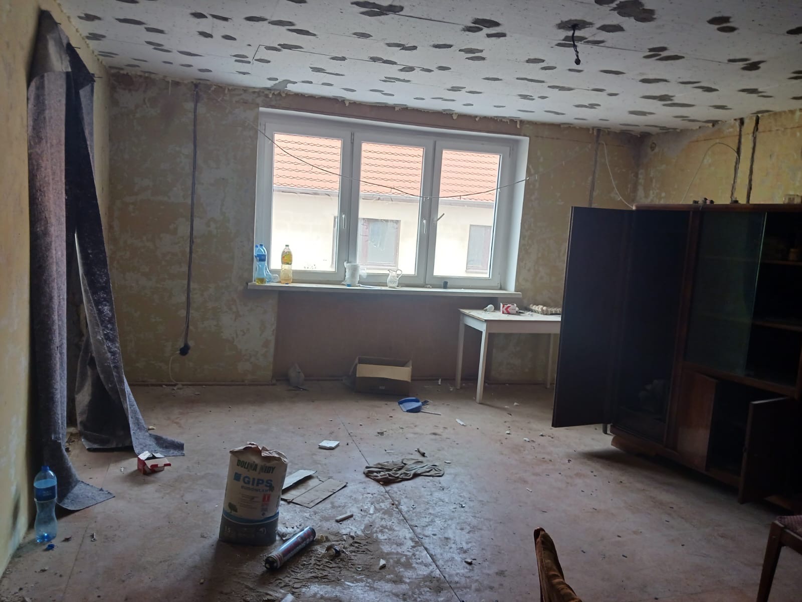 Pokój w trakcie remontu: ściany przygotowane pod prace wykończeniowe, widoczny gips budowlany, okno z widokiem na dach sąsiedniego budynku, częściowo zakryte meble, ślady po niedokończonych pracach...