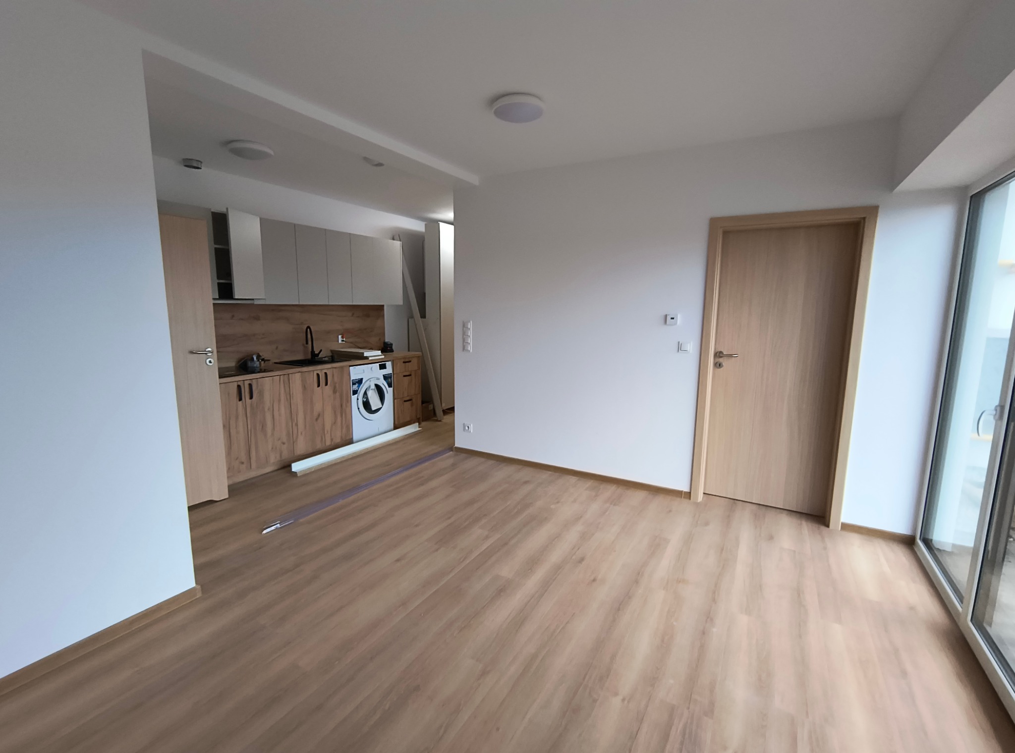 Jasne wnętrze mieszkania po remoncie, widoczne panele podłogowe w odcieniach drewna, białe ściany, drewniane drzwi wewnętrzne oraz zabudowa kuchenna z drewnianymi frontami i sprzętem AGD, duże okno...