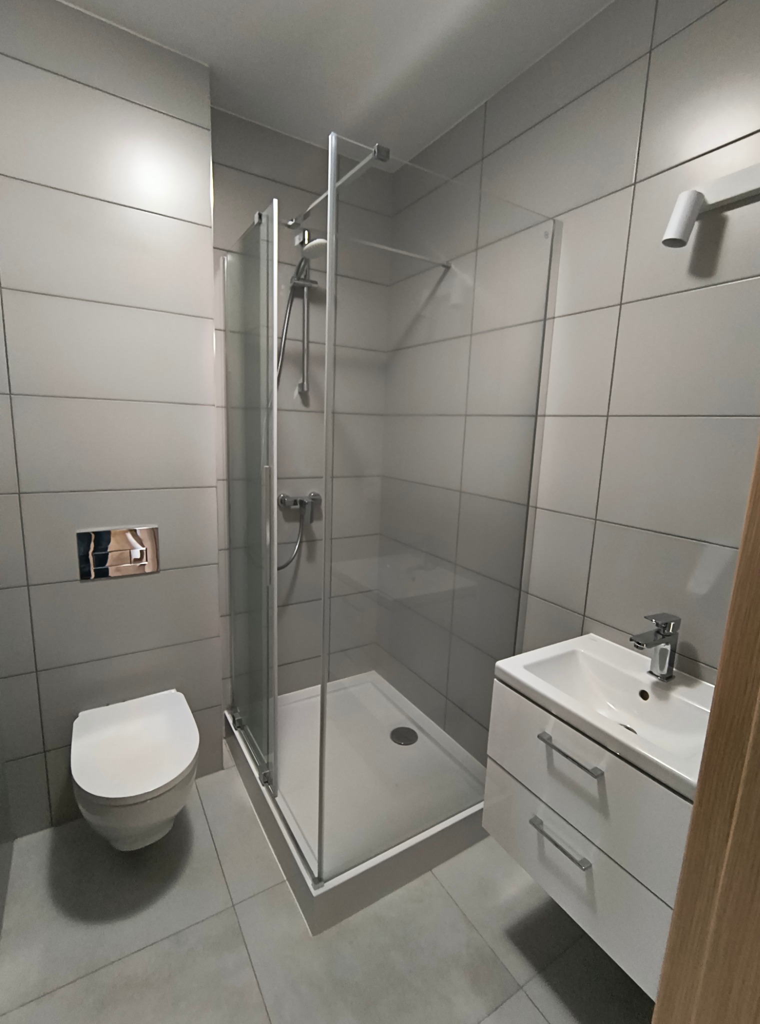 Nowoczesna łazienka wykończona szarymi, kwadratowymi płytkami, z kabiną prysznicową ze szklanymi drzwiami, podwieszaną toaletą i białą szafką z umywalką.