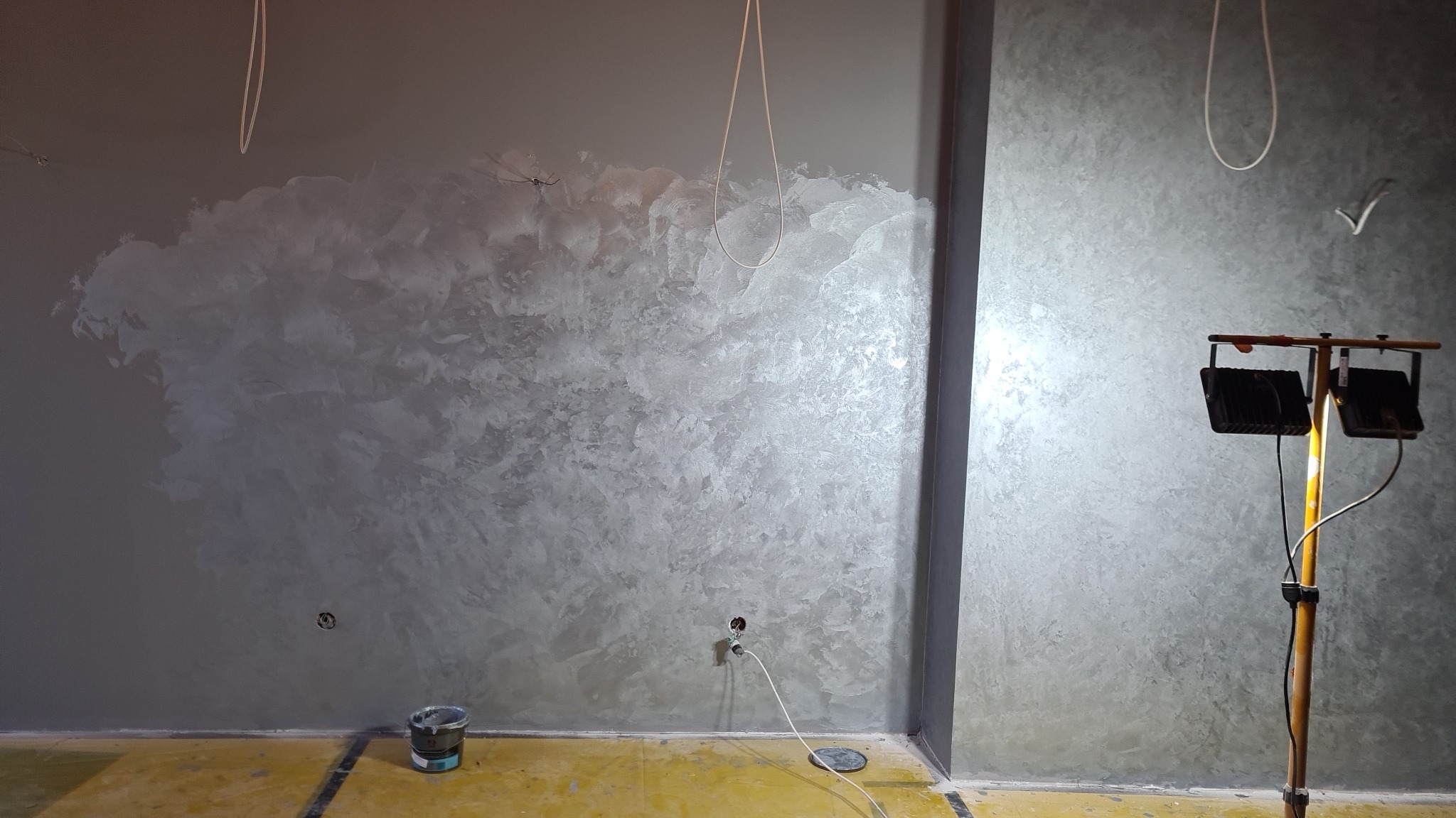 Ściana w szarym odcieniu z dekoracyjną, częściowo nałożoną strukturą, oświetlona lampą budowlaną, widoczny kabel wystający ze ściany i wiadro z farbą na podłodze zabezpieczonej folią.