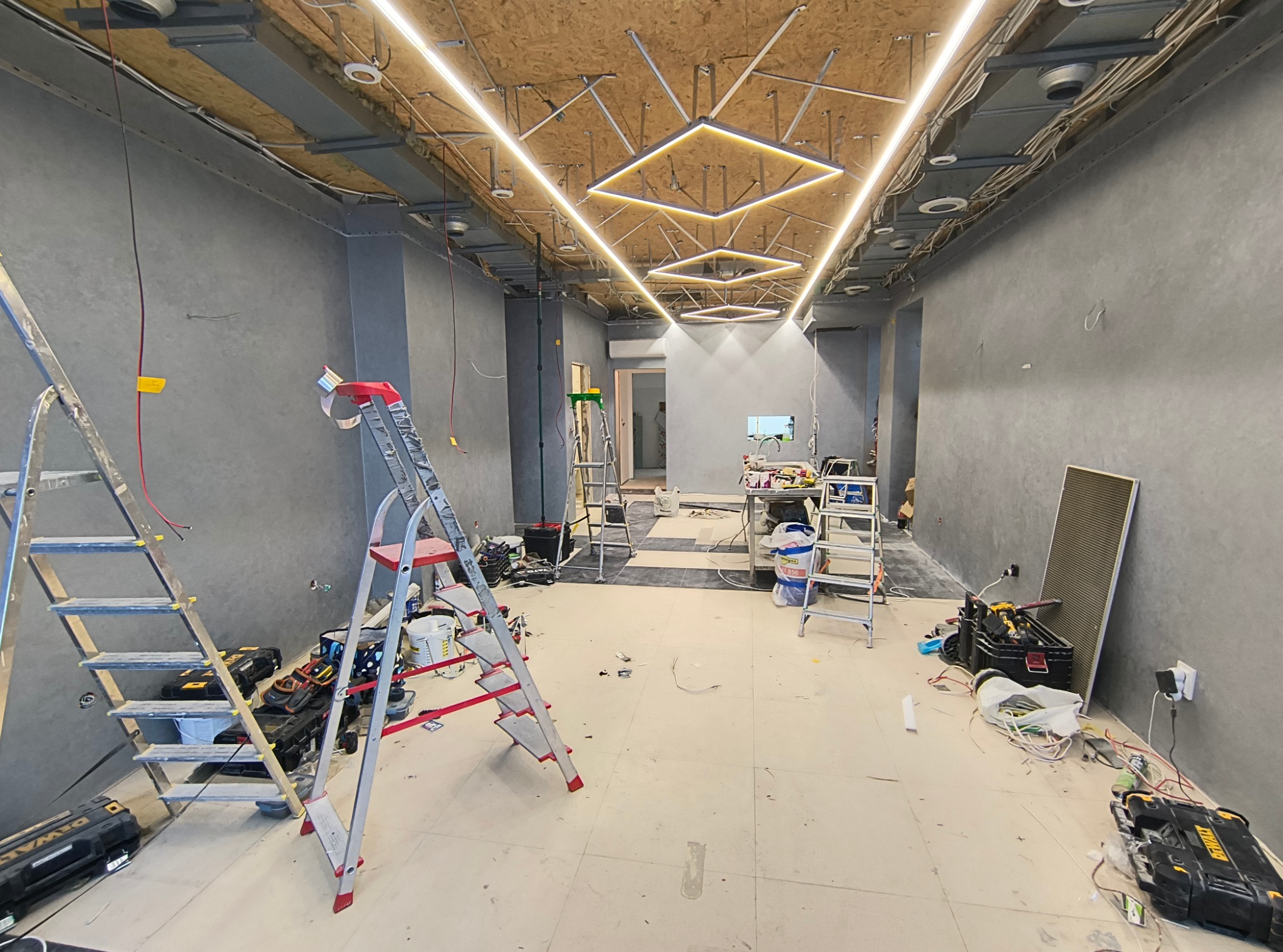 Wnętrze w trakcie prac wykończeniowych: szare ściany, sufit z odsłoniętą konstrukcją i zamontowanym oświetleniem LED w kształcie kwadratów, dwie aluminiowe drabiny, narzędzia i materiały budowlane...