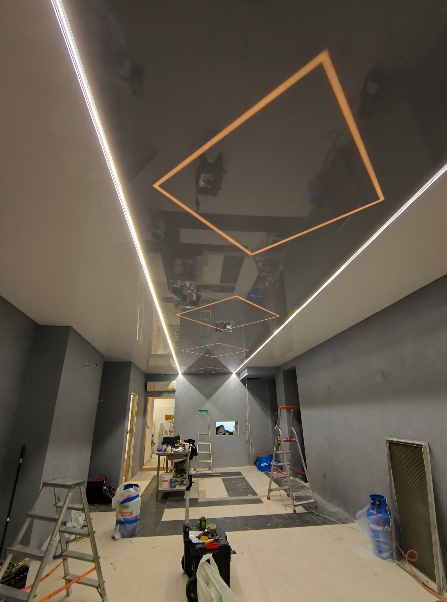 Sufit napinany z podświetleniem LED w kształcie kwadratów w trakcie montażu w pomieszczeniu w Zielonej Górze, widoczne drabiny, narzędzia i materiały budowlane.