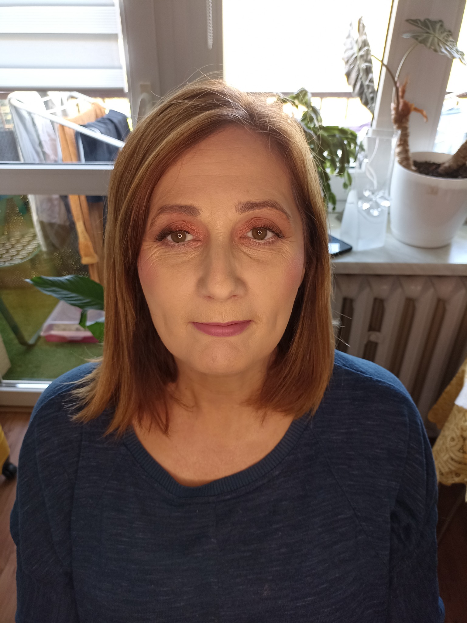 Portret kobiety z delikatnym makijażem oczu w odcieniach brzoskwini i ustami w kolorze mauve, sfotografowanej w domowym otoczeniu z widocznymi roślinami i suszarką na pranie w tle.