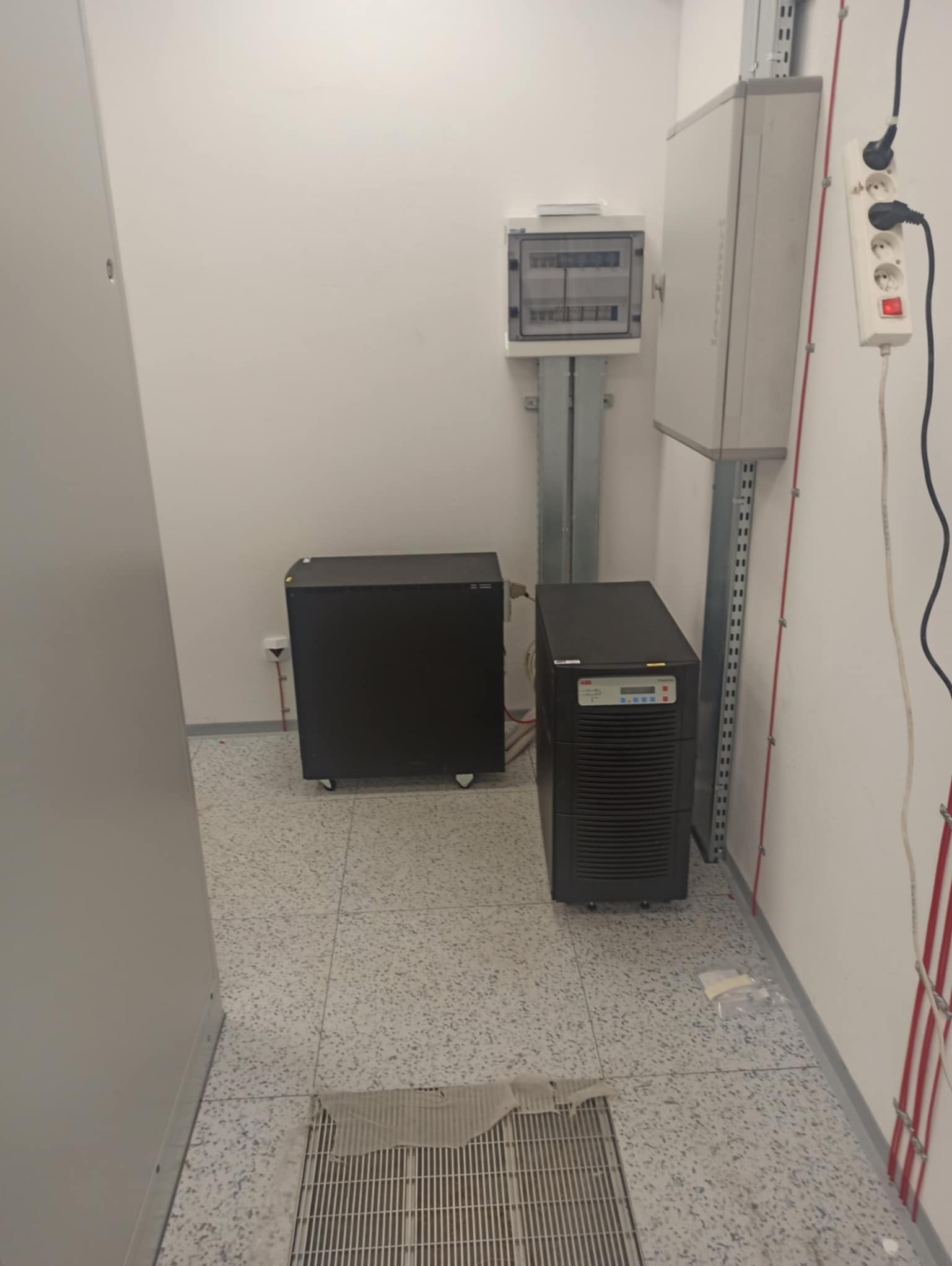 Zestaw zasilania awaryjnego UPS w serwerowni. Dwa urządzenia UPS, rozdzielnica elektryczna, listwa zasilająca. Białe ściany, szara podłoga w kropki.