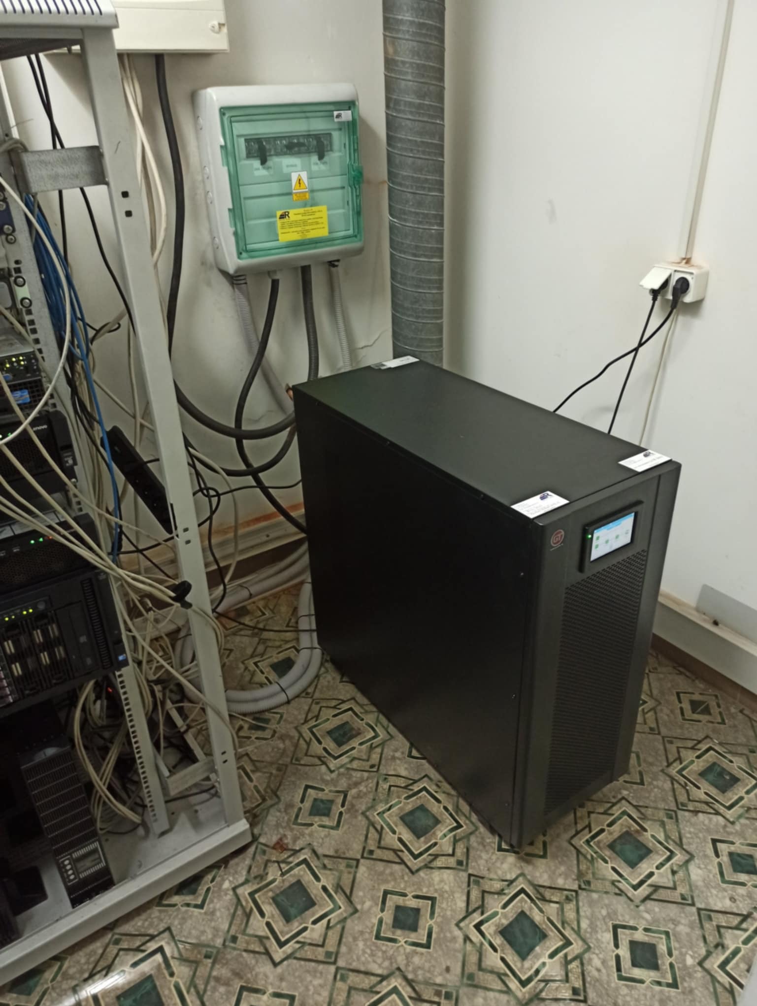 Czarna obudowa UPS z ekranem dotykowym, obok szafy serwerowej i skrzynki elektrycznej na wzorzystej podłodze. Widoczne kable i przewody.