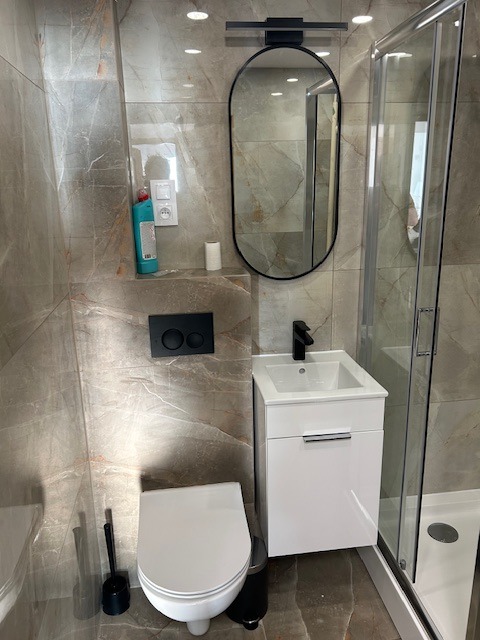 Nowoczesna łazienka z marmurowymi płytkami, owalnym lustrem z czarną ramą, białą szafką umywalkową i kabiną prysznicową. Widoczny kompakt WC i czarne akcenty.