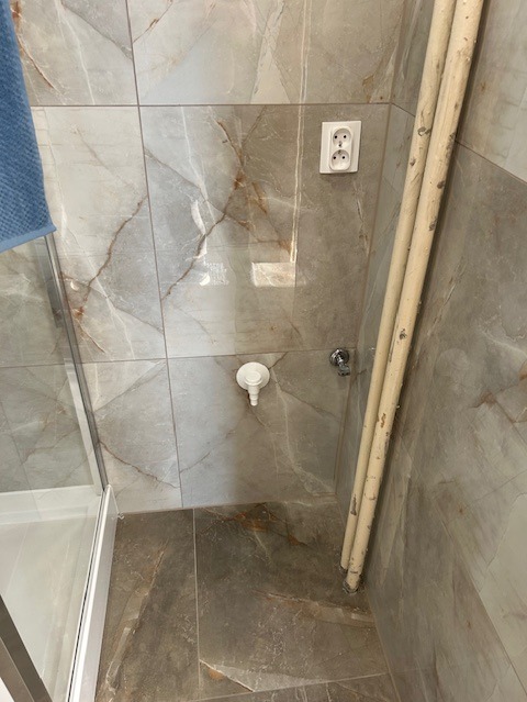 Wykończona łazienka z marmurowymi płytkami, widoczne rury grzewcze, odpływ liniowy i gniazdko elektryczne. Estetyczne i funkcjonalne rozwiązanie.