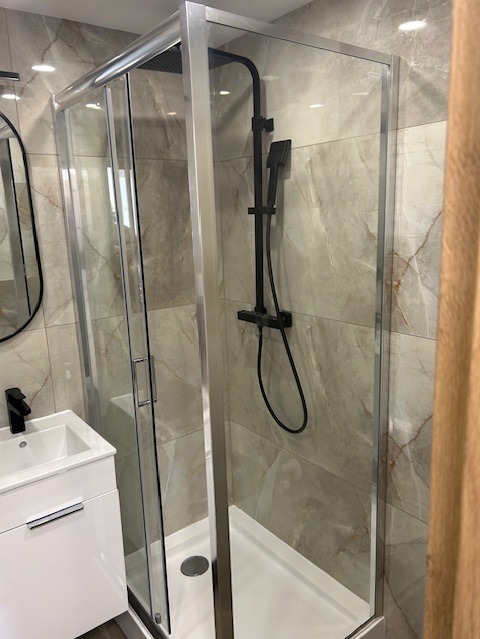 Nowoczesna kabina prysznicowa z czarną armaturą na tle marmurowych płytek. Widoczna umywalka z szafką. Minimalistyczny design, eleganckie wykończenie.