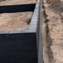 Wykopane fundamenty domu z widoczną izolacją przeciwwilgociową, teren budowy z wykopaną ziemią i roślinnością w tle.