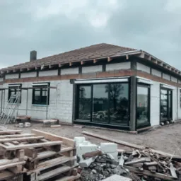 Niedokończony budynek z białych bloczków z dużymi oknami, widoczne drewniane elementy konstrukcji dachu i rusztowanie, palety z materiałami budowlanymi na pierwszym planie, pochmurne niebo w tle.