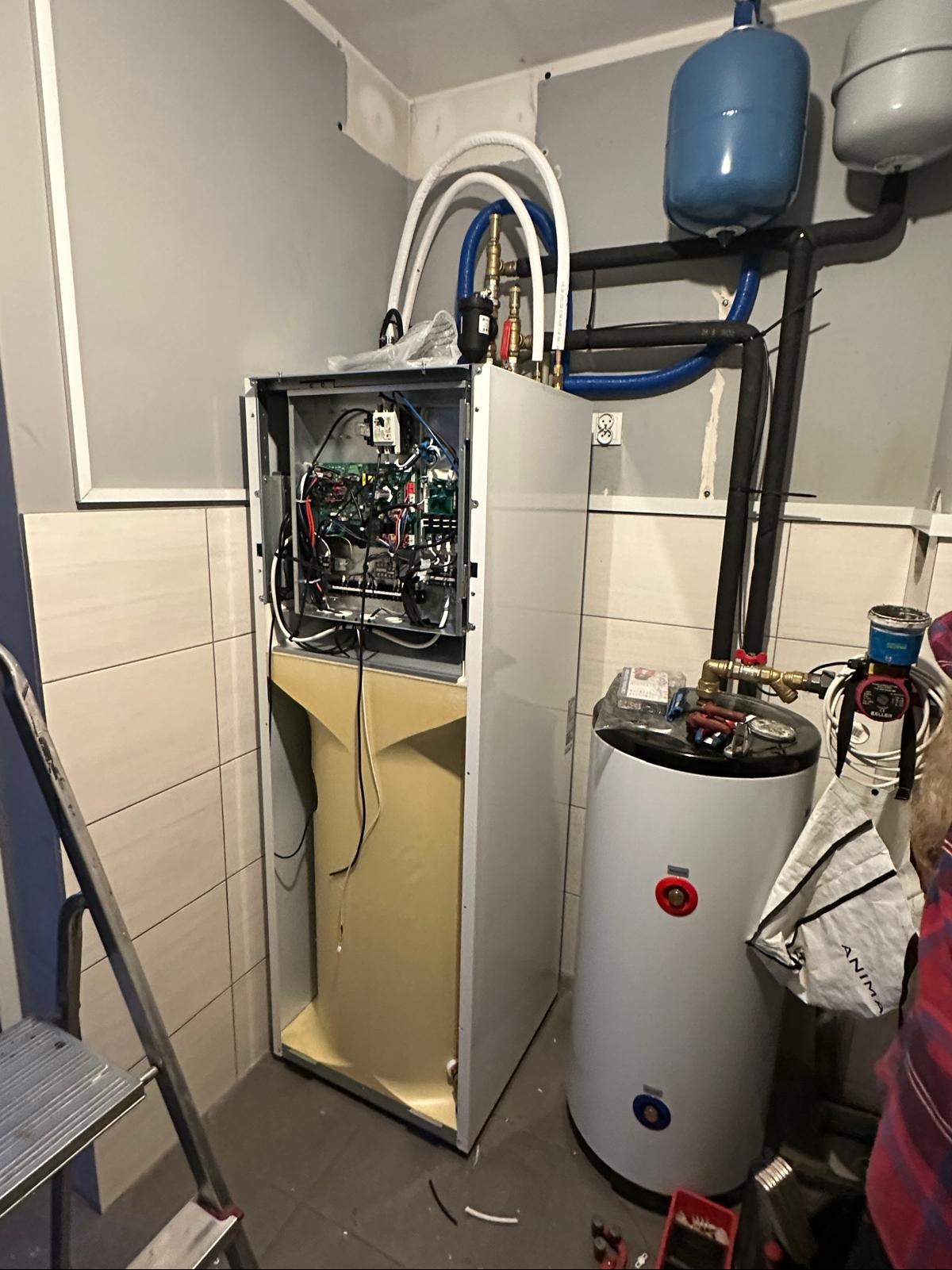 Otwarta obudowa kotła grzewczego z widocznymi przewodami i elektroniką, obok biały zasobnik ciepłej wody z osprzętem w kotłowni z drabinką.