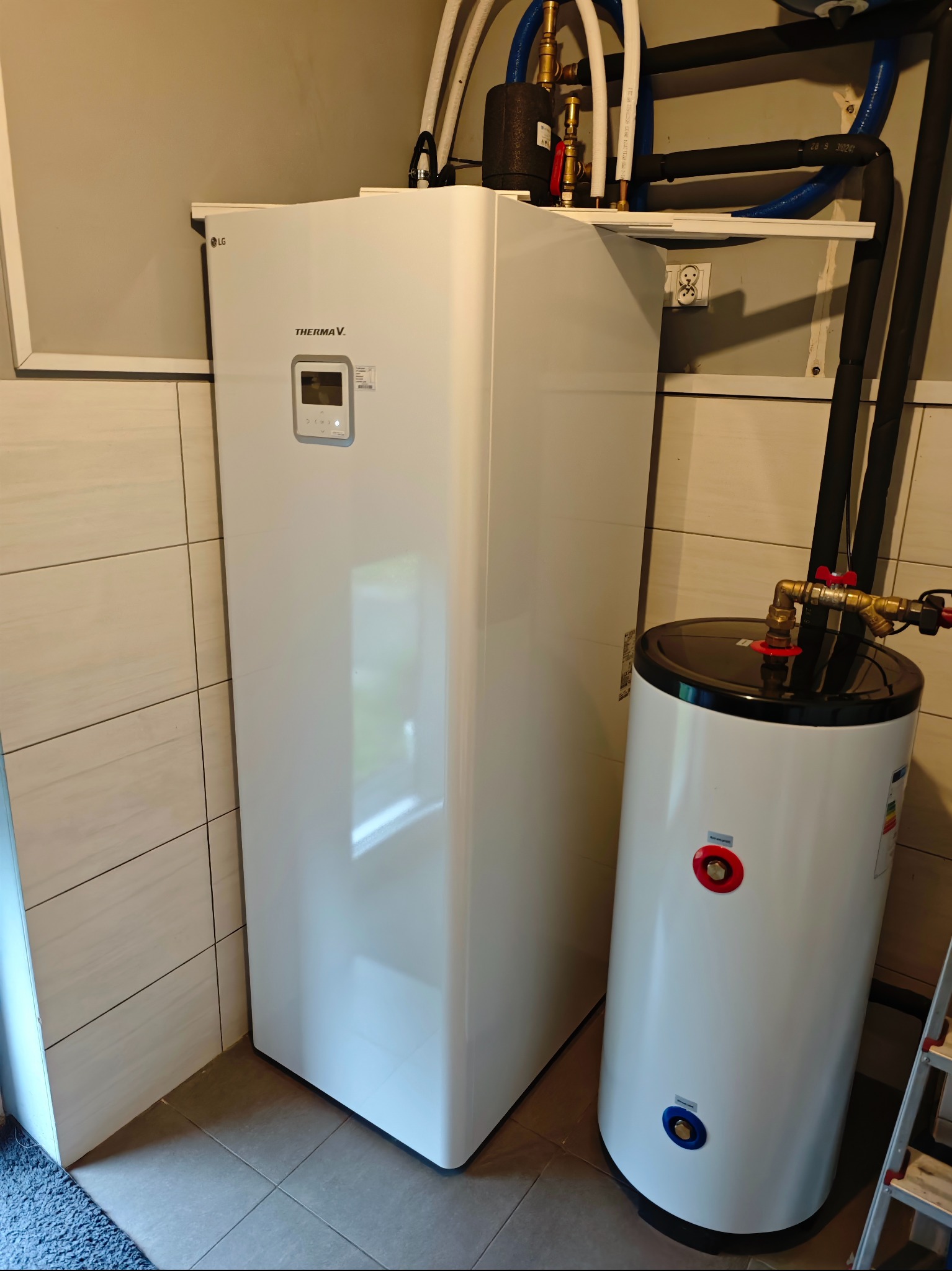 Biała pompa ciepła LG Therma V z zbiornikiem, widoczne rury instalacyjne i zawory, zamontowana w pomieszczeniu z jasnymi płytkami.