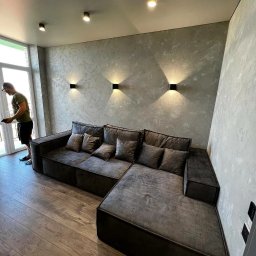 D&L remont - Szary narożnik z poduszkami w salonie z drewnianą podłogą i ścianami pokrytymi dekoracyjną strukturą, oświetlonymi trzema czarnymi kinkietami.