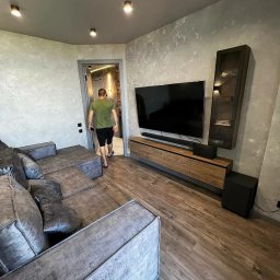 D&L remont - Nowoczesny salon z szarą narożną sofą, drewnianą podłogą i ścianami wykończonymi w stylu betonu architektonicznego; w tle widoczny mężczyzna wchodzący do pokoju.