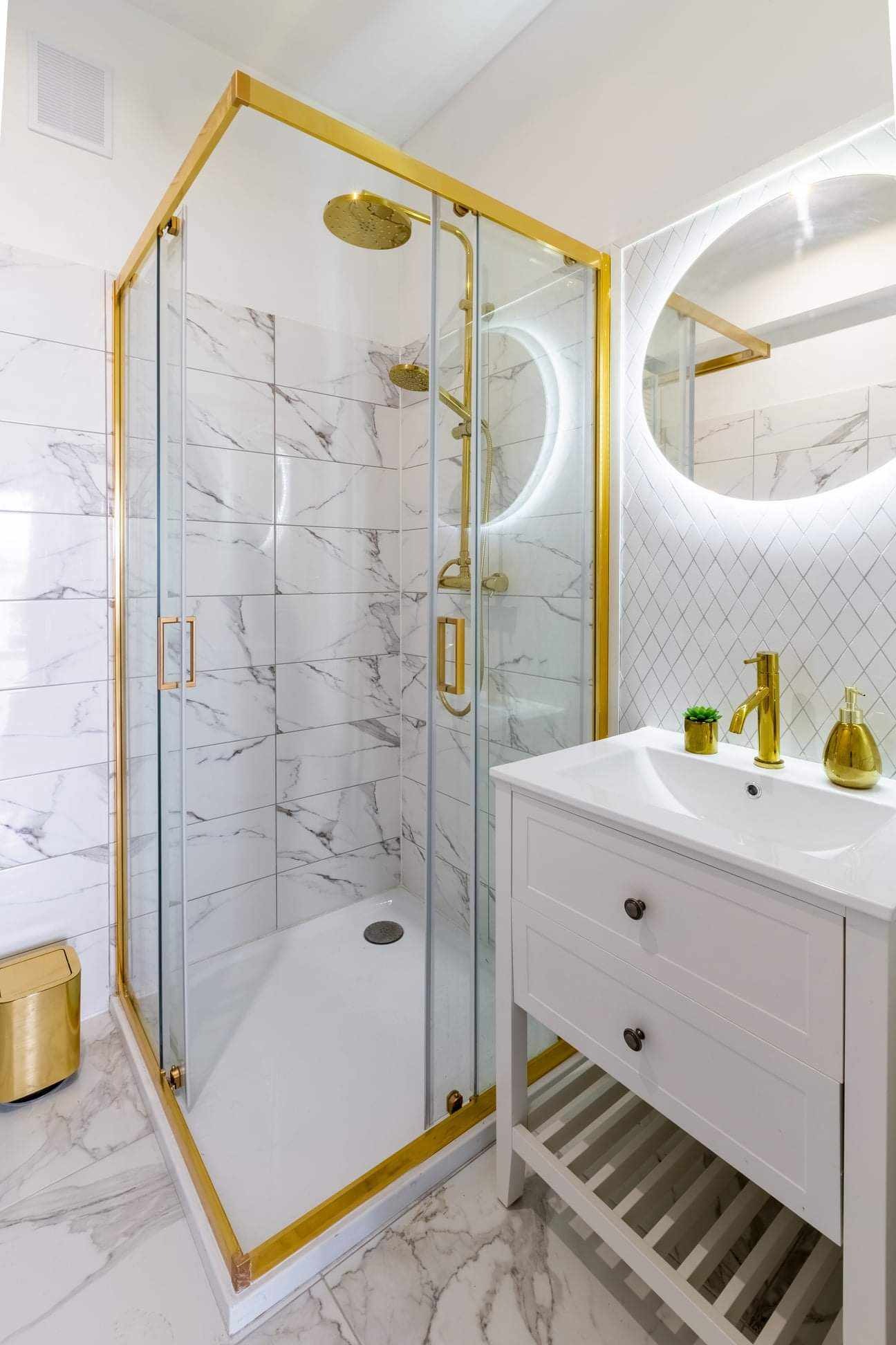 Nowoczesna łazienka z kabiną prysznicową w złotej ramie, marmurowe płytki na ścianach i podłodze, biała umywalka z szafką i okrągłym lustrem z podświetleniem LED.