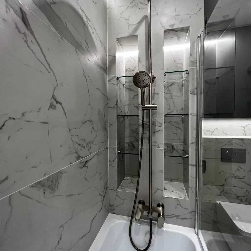 Wykończona łazienka z płytkami imitującymi marmur, zabudowana wanna z chromowaną baterią i deszczownicą, w ścianie wnęki z szklanymi półkami i podświetleniem LED.