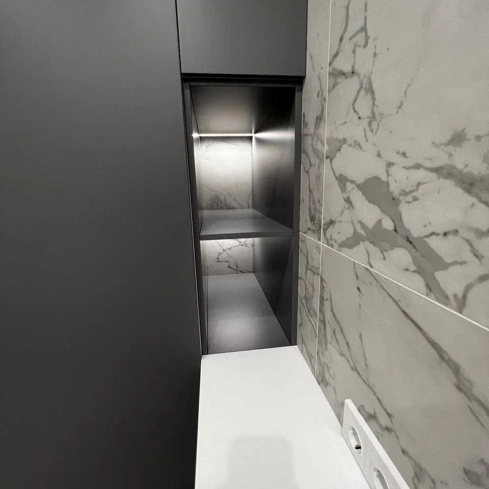 Nowoczesna łazienka z szarymi płytkami imitującymi marmur, czarną szafką w zabudowie i podświetlaną wnęką z półkami.