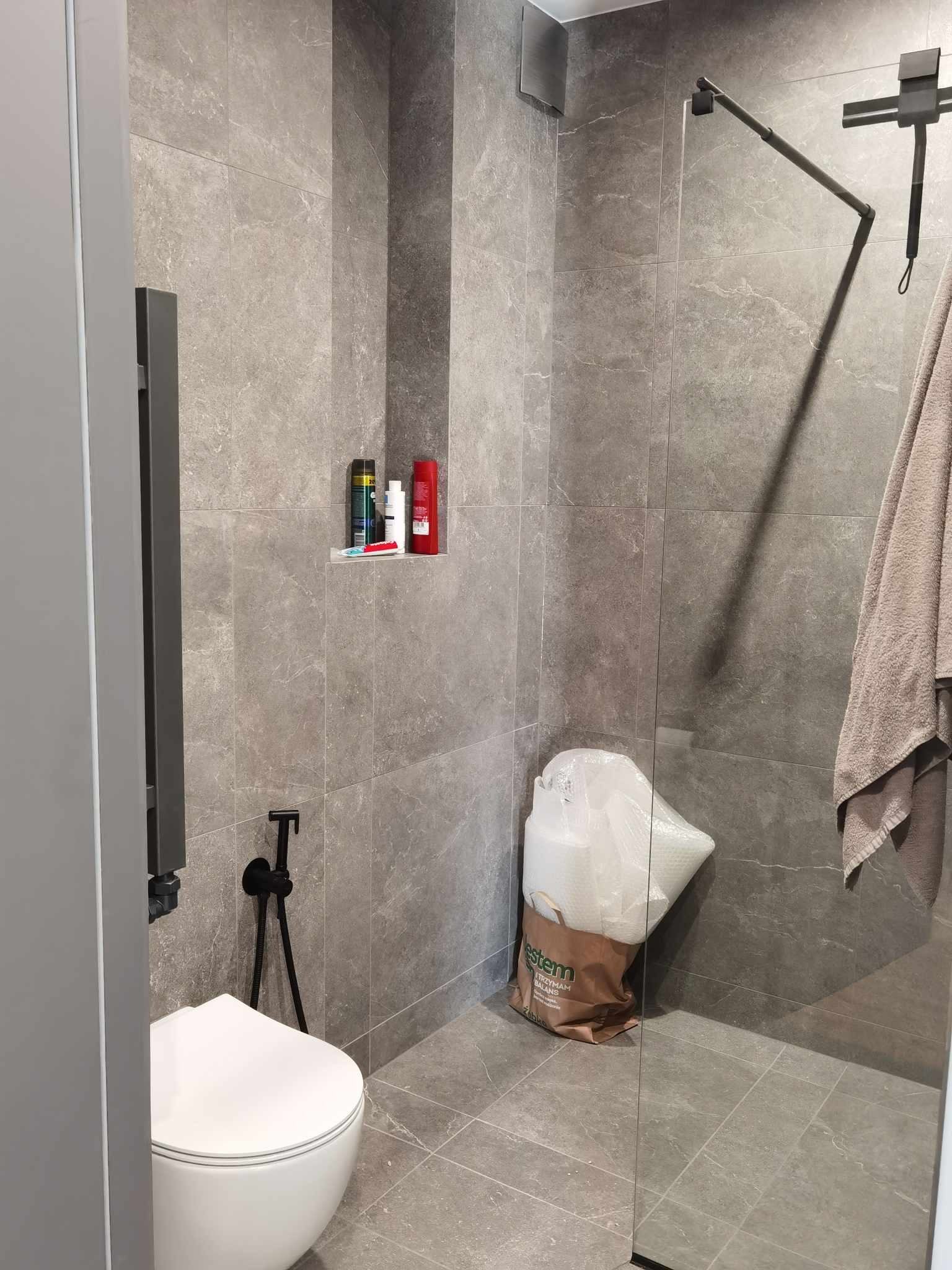 Szara łazienka z kabiną prysznicową typu walk-in, wiszącą toaletą, grzejnikiem drabinkowym i workiem z materiałami budowlanymi w rogu.