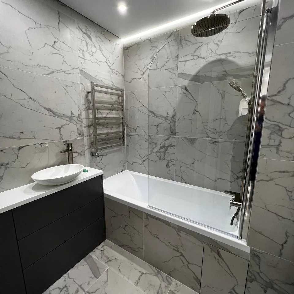 Nowoczesna łazienka z marmurowymi płytkami, owalną umywalką nablatową, grzejnikiem drabinkowym i kabiną nawannową z chromowanymi profilami, oświetlona górnym światłem punktowym i paskiem LED.