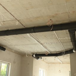 Klimbrand Adrian Kaflik - Widok instalacji rekuperacji z czarnymi rurami pod betonowym sufitem w surowym wnętrzu budynku w trakcie wykończenia, widoczne kable elektryczne i okno.