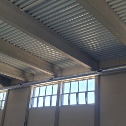 Klimbrand Adrian Kaflik - Szary, metalowy kanał wentylacyjny biegnący pod sufitem hali przemysłowej z widocznymi oknami i betonowymi belkami stropowymi.