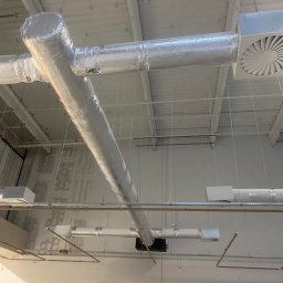 Klimbrand Adrian Kaflik - Widok z dołu na system kanałów wentylacyjnych z izolacją aluminiową, kratki wentylacyjne i częściowo widoczne podnośniki koszowe w jasnym pomieszczeniu przemysłowym.