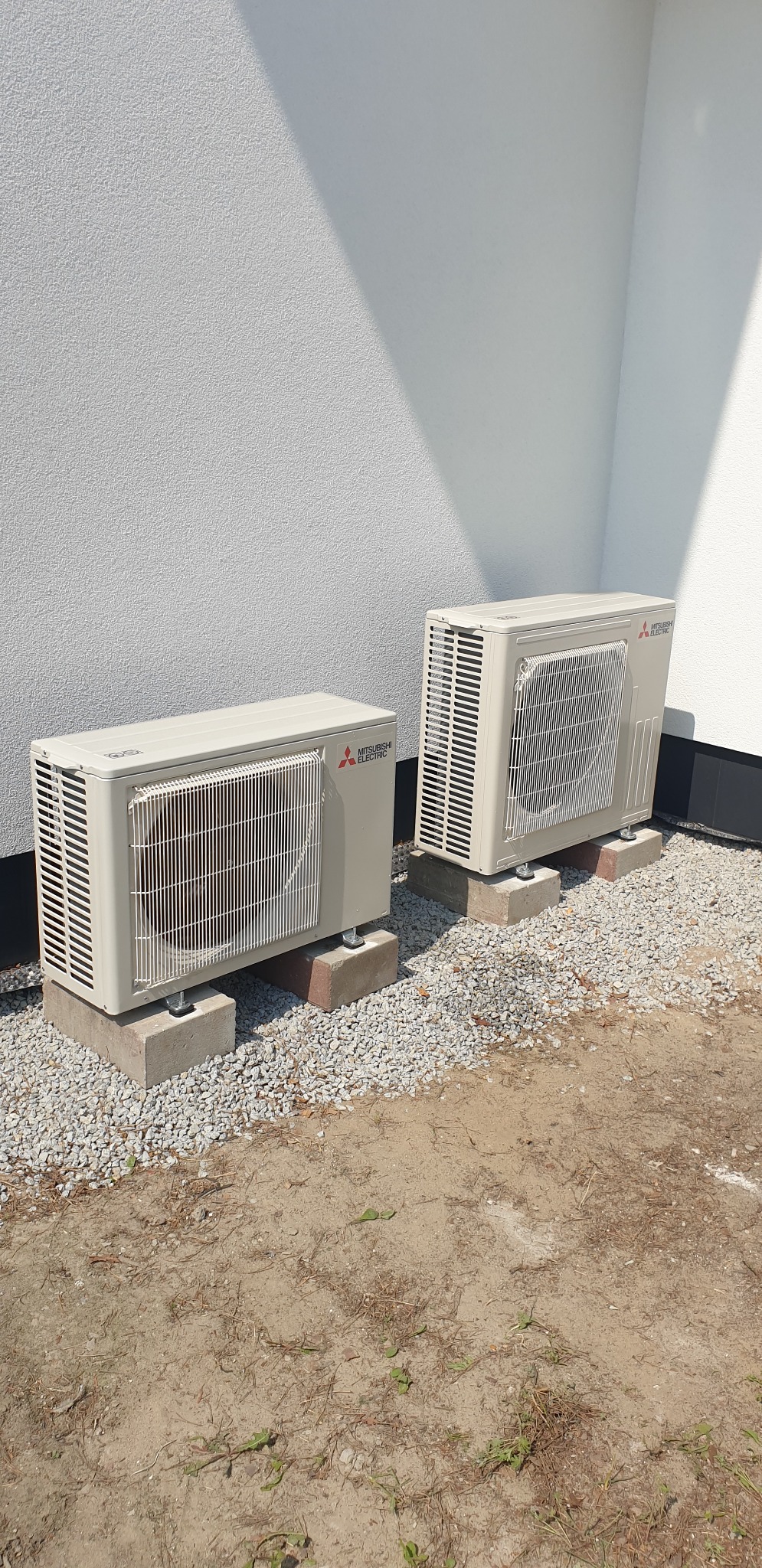 Dwie jednostki zewnętrzne klimatyzacji Mitsubishi Electric ustawione na betonowych podstawach, otoczone drobnym żwirem przy białej ścianie budynku.