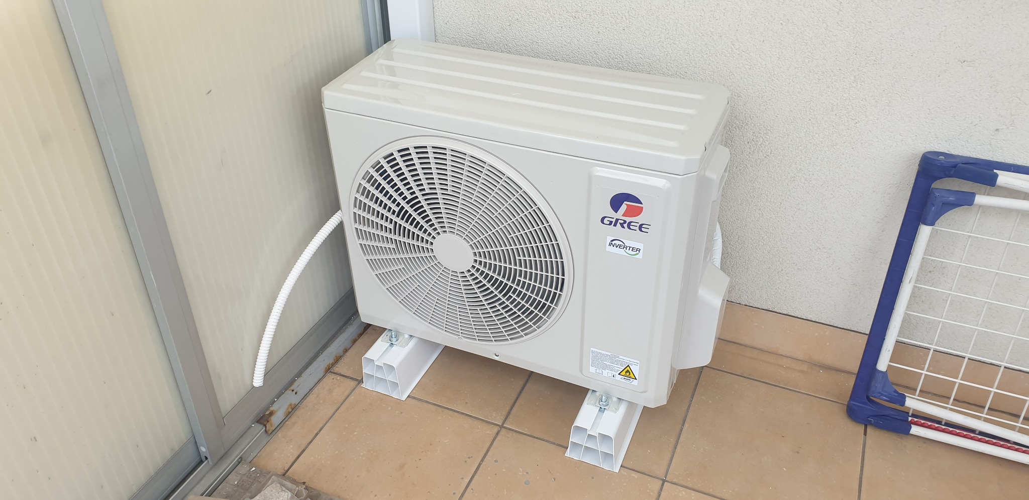 Zewnętrzna jednostka klimatyzacji marki Gree zamontowana na białych wspornikach na brązowych płytkach tarasowych, z fragmentem suszarki do ubrań w tle.