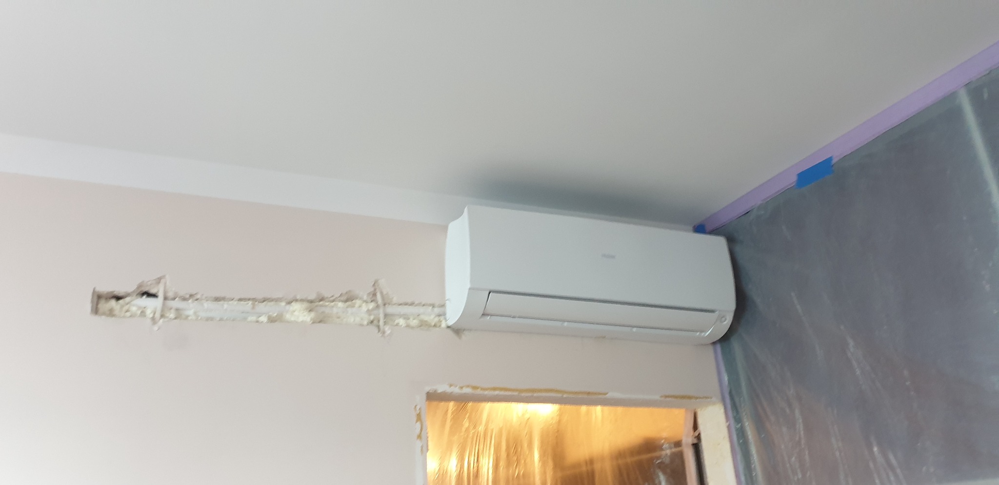 Biały klimatyzator Haier zamontowany na ścianie w trakcie instalacji, widoczne przewody w ścianie zabezpieczone pianką montażową, ściana częściowo zabezpieczona folią malarską.