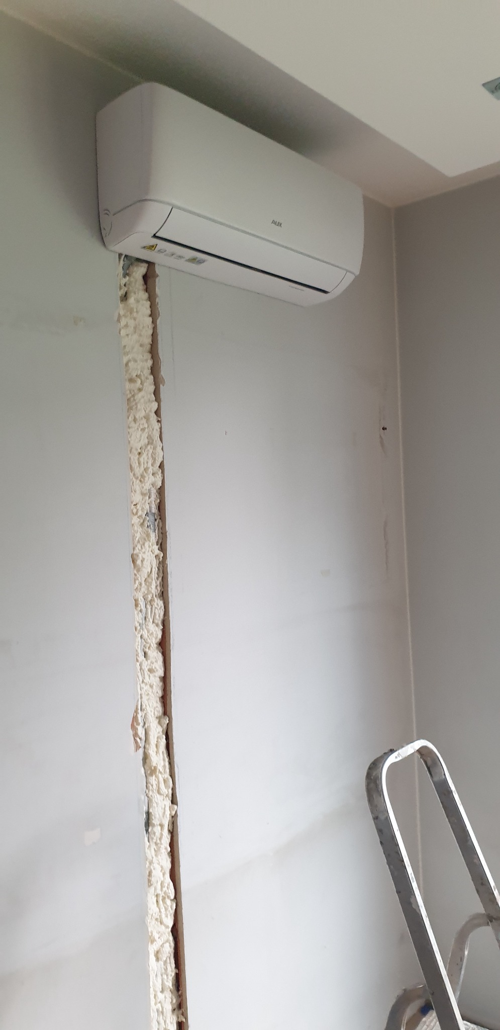 Biały klimatyzator AUX zamontowany na ścianie, widoczna szczelina wypełniona pianką montażową oraz fragment drabiny malarskiej.