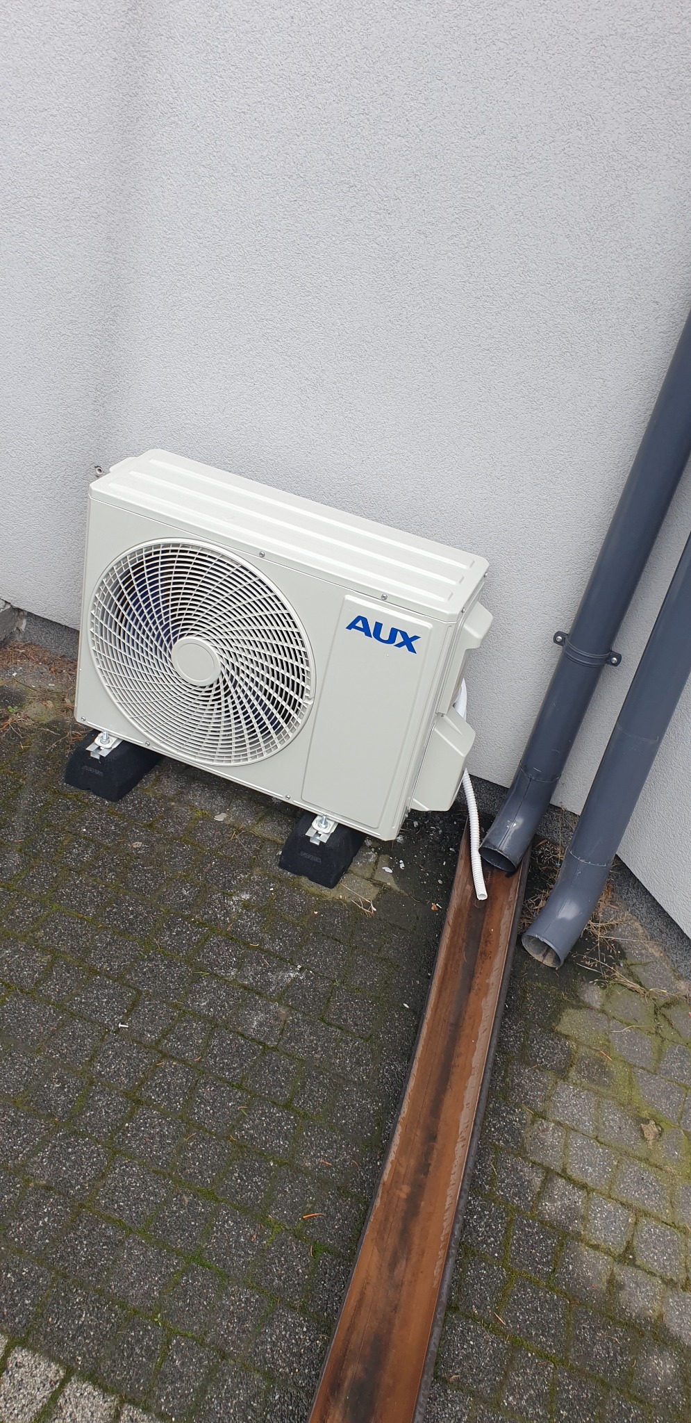 Zewnętrzna jednostka klimatyzacji marki AUX zamontowana na gumowych podkładkach przy ścianie budynku, obok rynien spustowych.