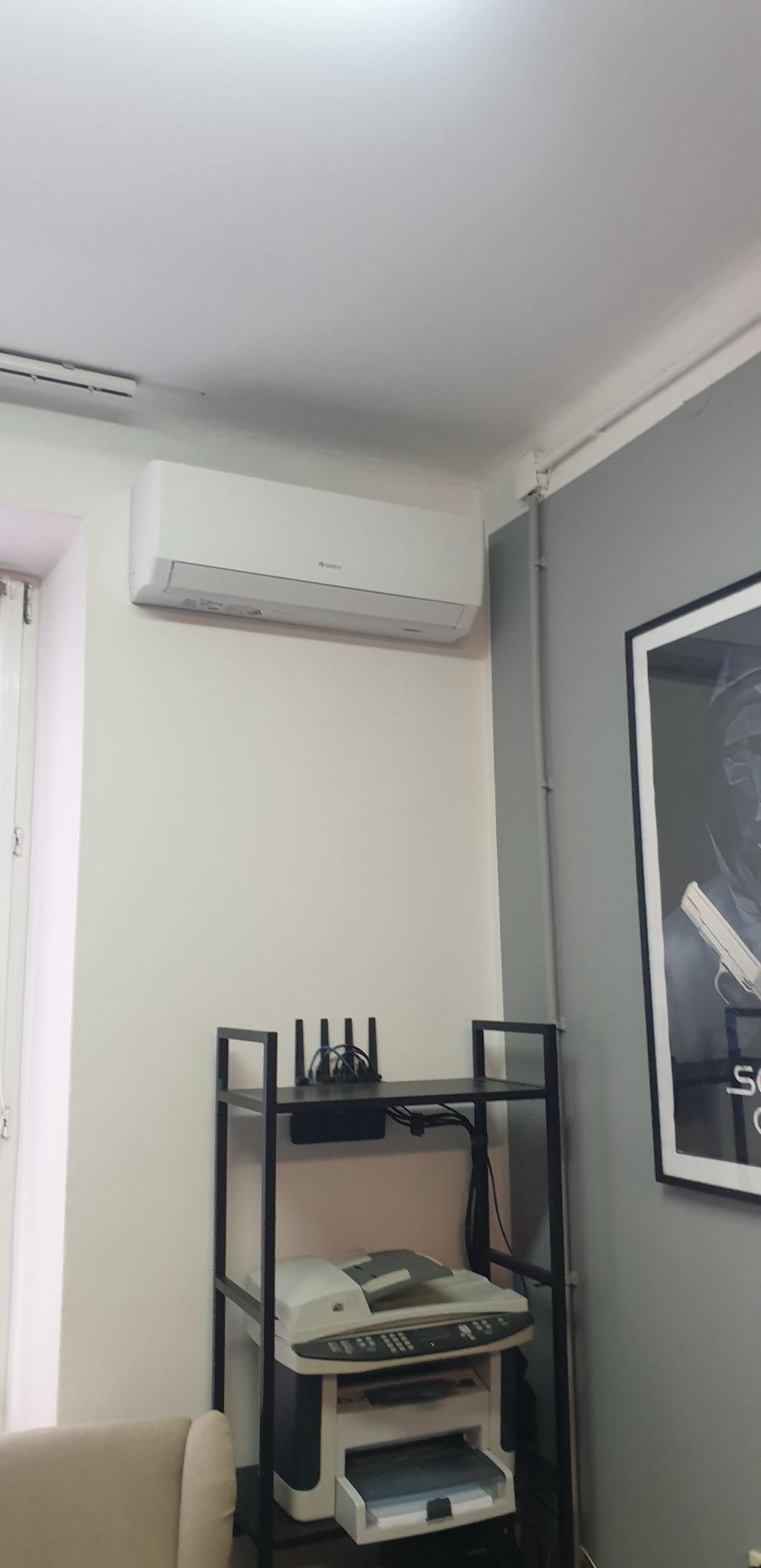 Biały klimatyzator ścienny marki Gree zamontowany w biurze, pod nim metalowy regał z routerem i drukarką wielofunkcyjną, widoczna część plakatu na szarej ścianie.