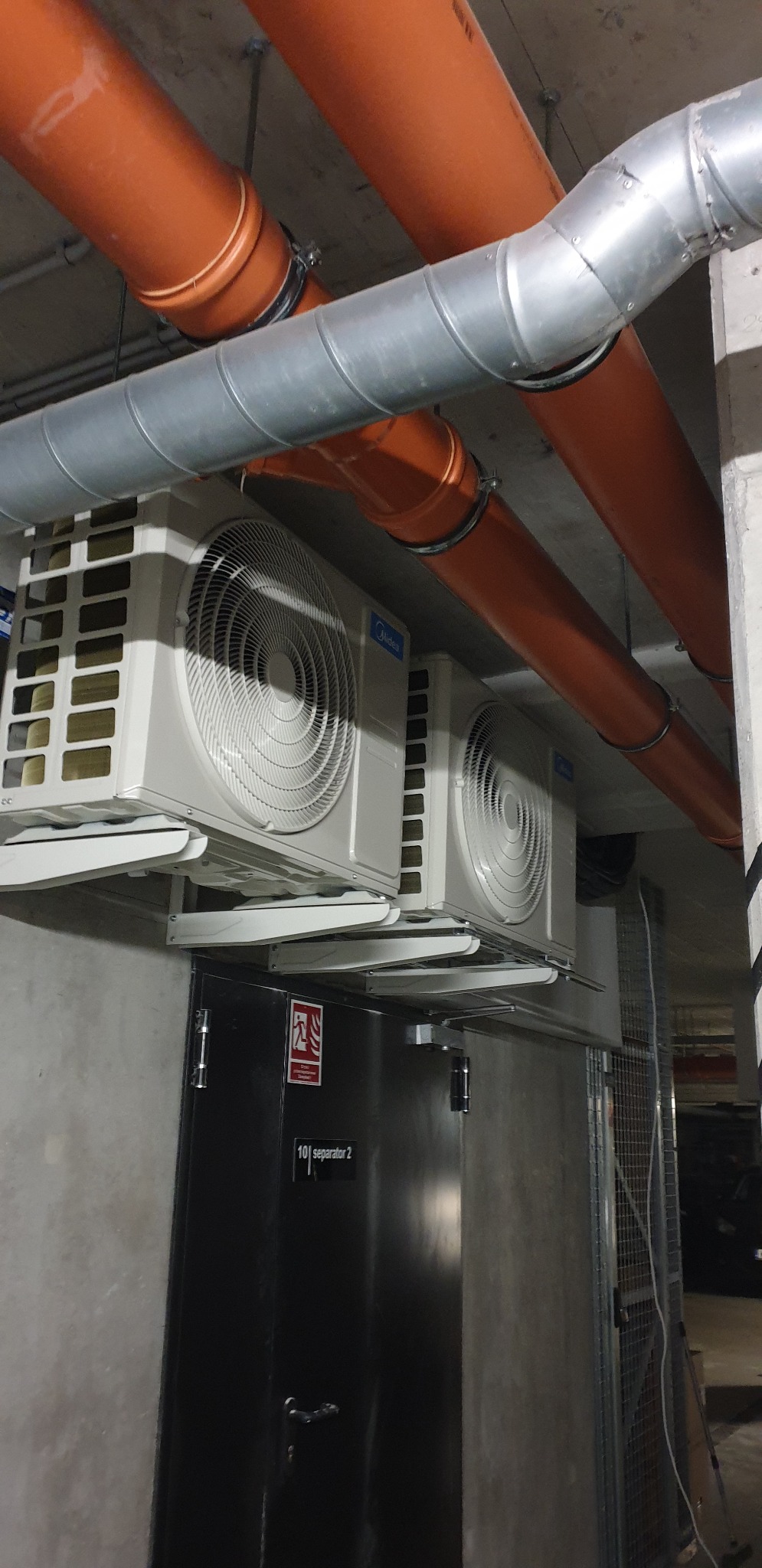 Dwa białe klimatyzatory Midea zamontowane pod sufitem w pomieszczeniu technicznym, widoczne rury wentylacyjne pomarańczowe i srebrne, oraz czarne drzwi z oznaczeniem separatora.