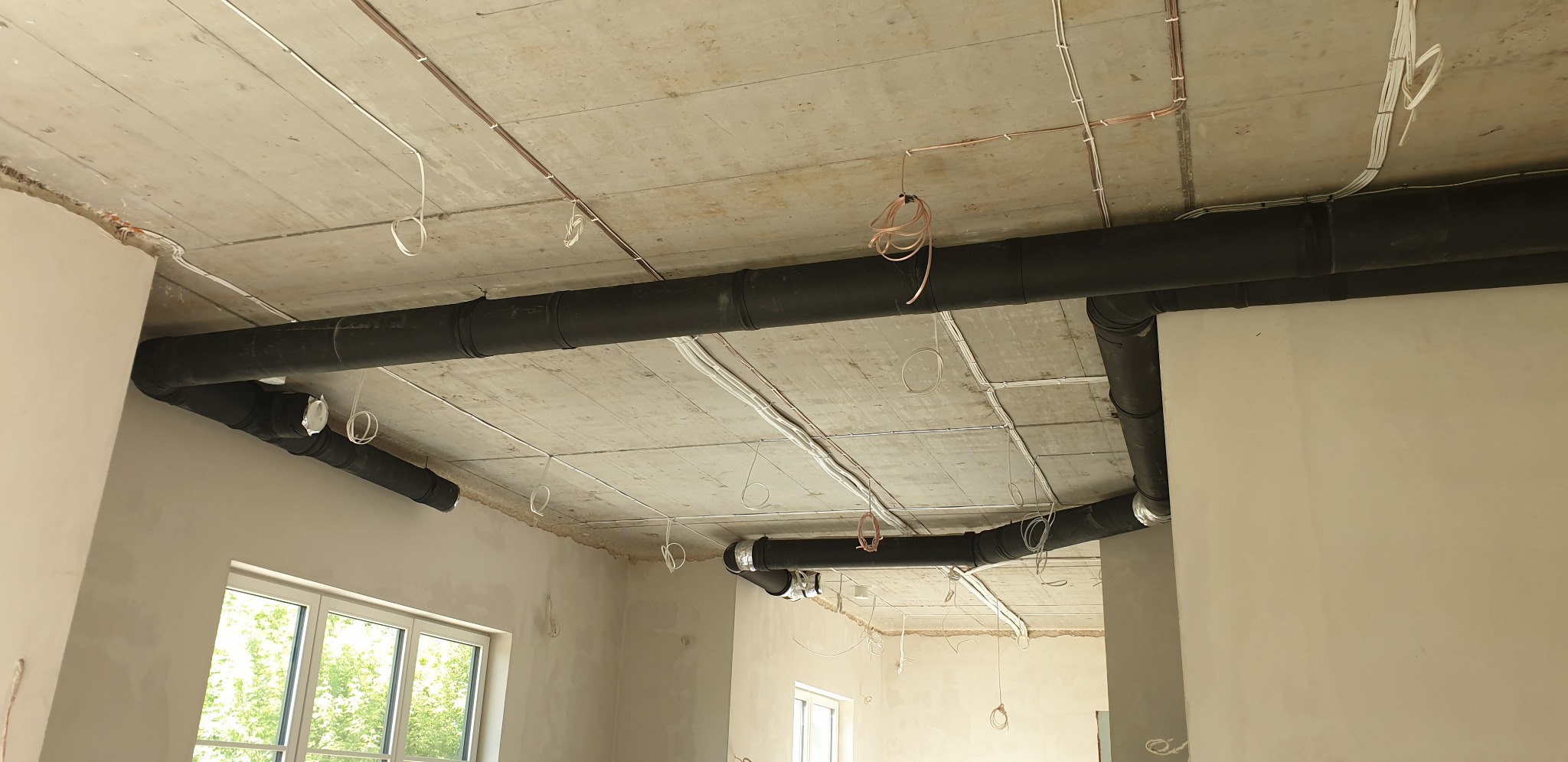 Widok instalacji rekuperacji z czarnymi rurami pod betonowym sufitem w surowym wnętrzu budynku w trakcie wykończenia, widoczne kable elektryczne i okno.