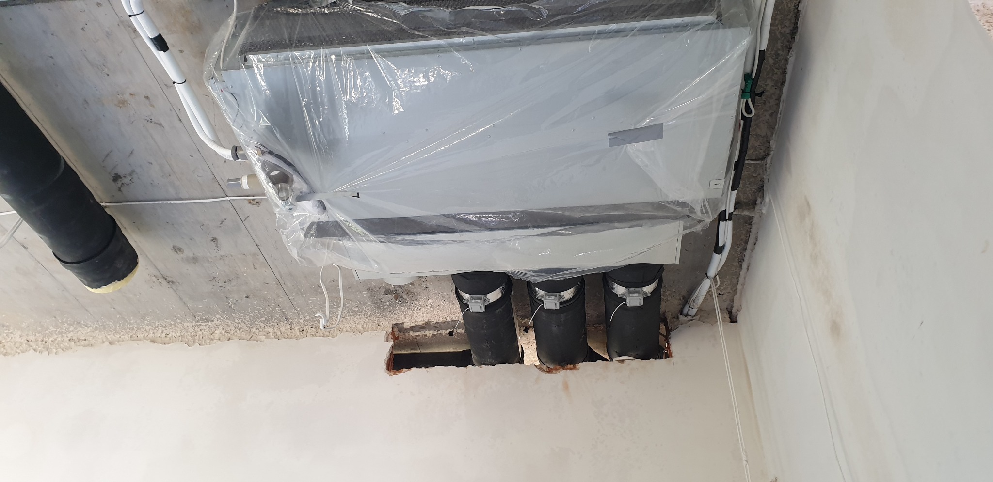 Kasetonowa jednostka klimatyzacyjna, owinięta folią ochronną, zamontowana w suficie z widocznymi przewodami i rurami wentylacyjnymi.