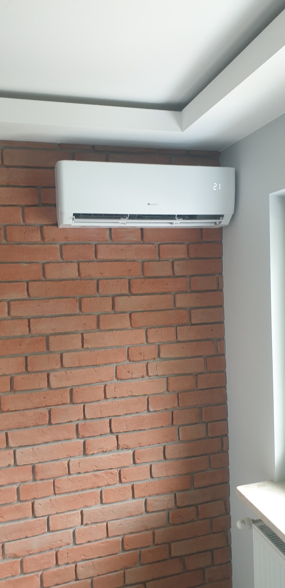 Biała jednostka klimatyzacyjna marki Gree zamontowana na ścianie z cegły w pomieszczeniu mieszkalnym, wyświetlająca temperaturę 21 stopni.