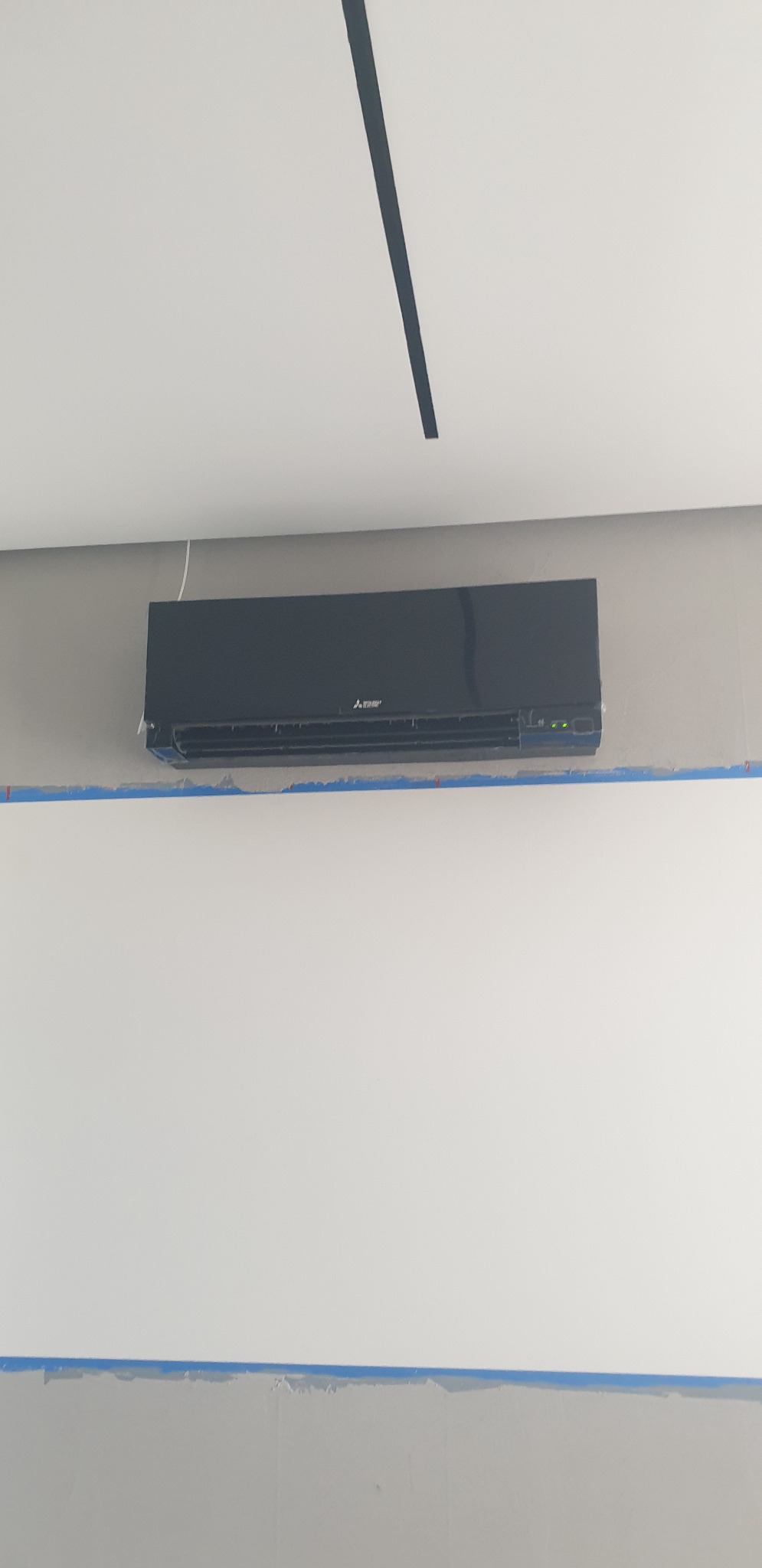 Czarny klimatyzator Mitsubishi zamontowany na szarej ścianie, zabezpieczonej taśmą malarską przed malowaniem.