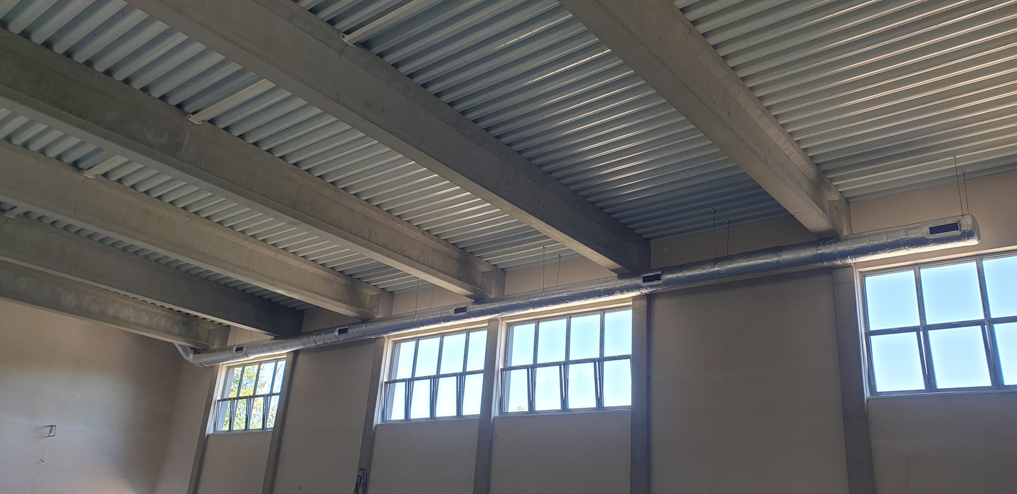 Szary, metalowy kanał wentylacyjny biegnący pod sufitem hali przemysłowej z widocznymi oknami i betonowymi belkami stropowymi.