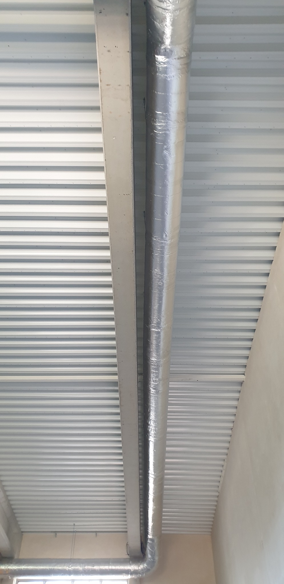 Widok z dołu na srebrną, izolowaną rurę wentylacyjną biegnącą wzdłuż metalowej belki pod sufitem z blachy trapezowej.