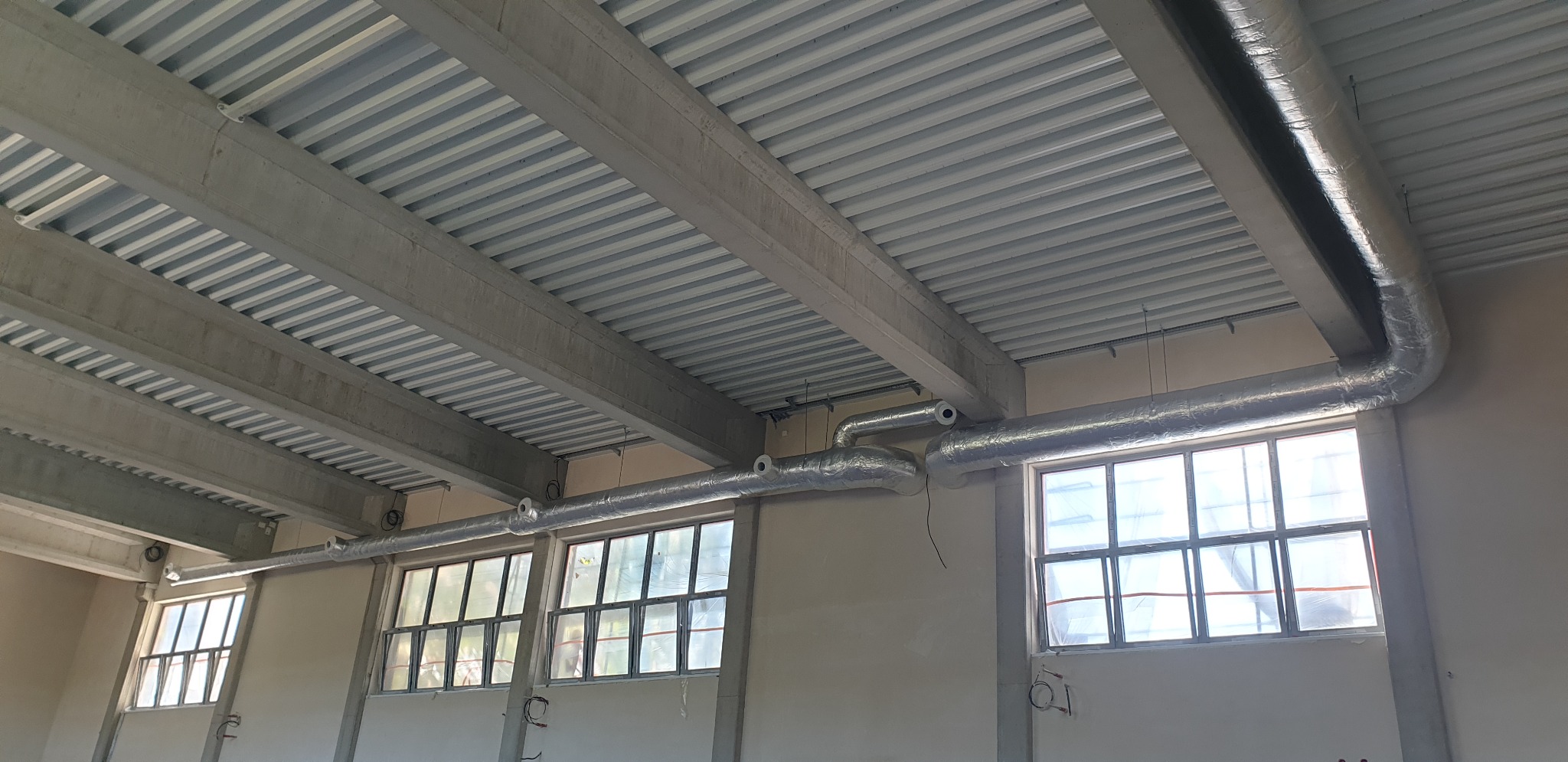 Instalacja srebrnych, izolowanych przewodów wentylacyjnych pod sufitem z blachy trapezowej w pomieszczeniu z oknami.