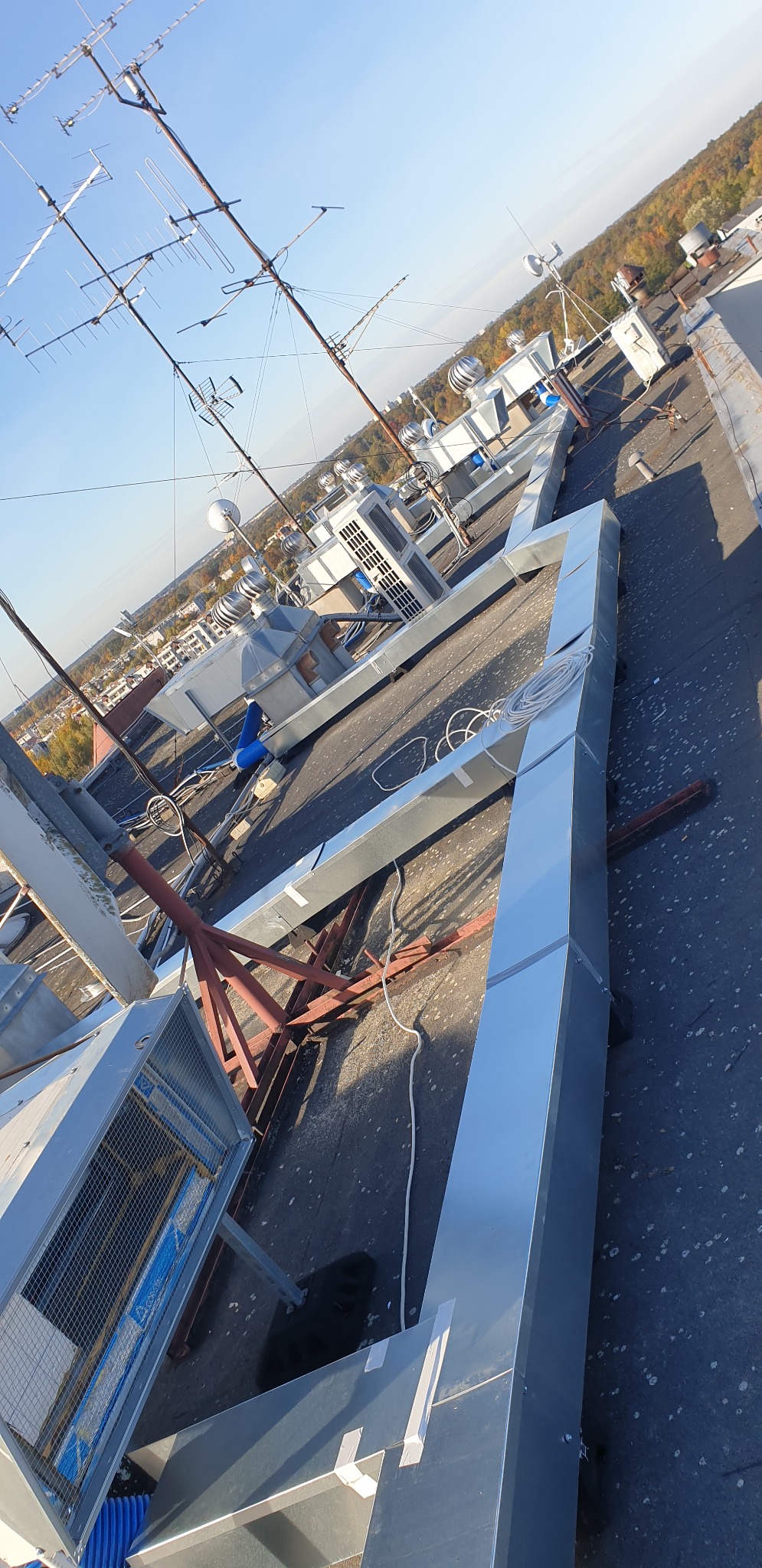 Widok z góry na instalację wentylacyjną na dachu budynku, z widocznymi kanałami wentylacyjnymi, centralami wentylacyjnymi i antenami na tle zabudowy miejskiej i drzew.