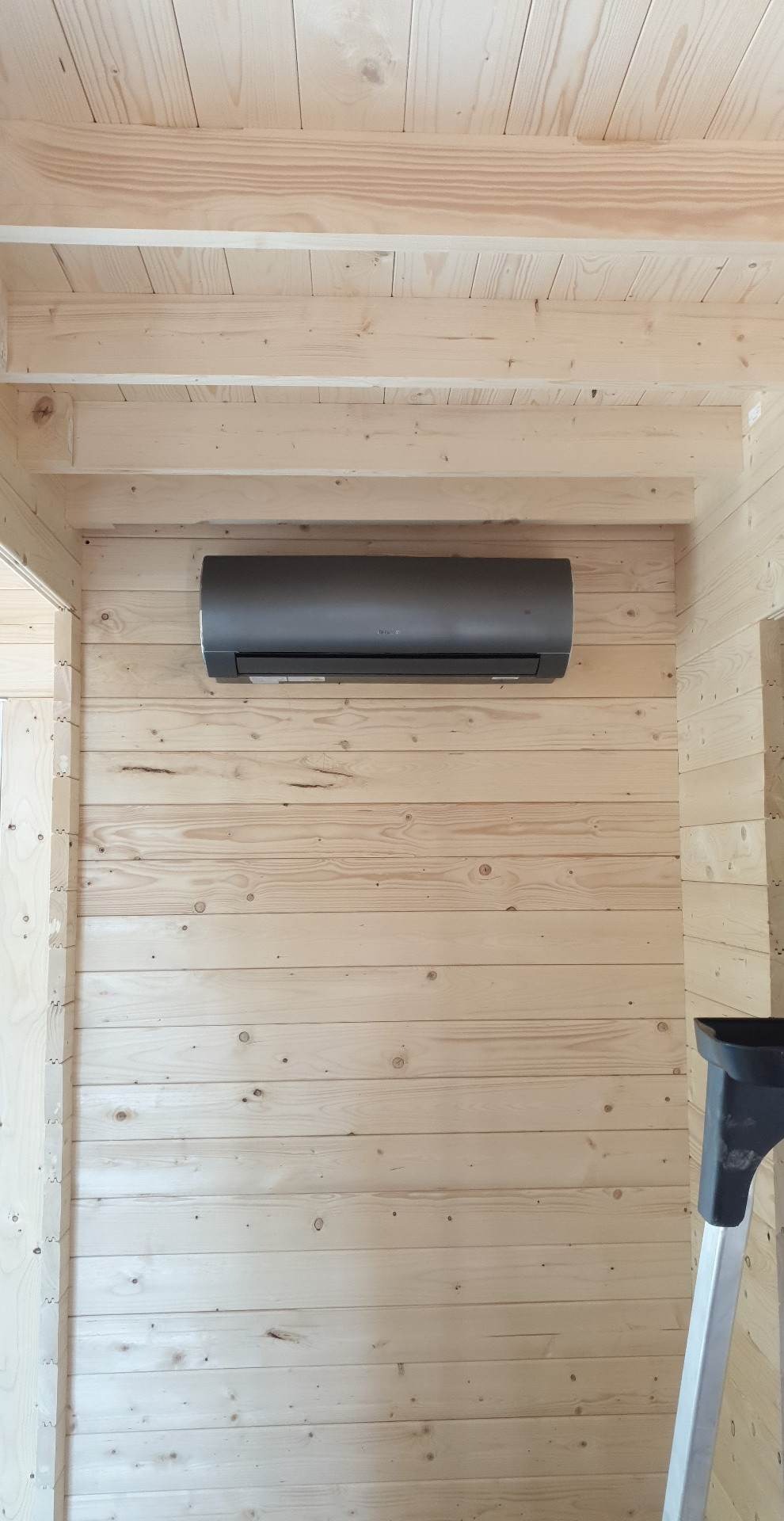 Szary klimatyzator zamontowany na ścianie z jasnego drewna w pomieszczeniu z drewnianym sufitem, widoczny fragment drabiny.