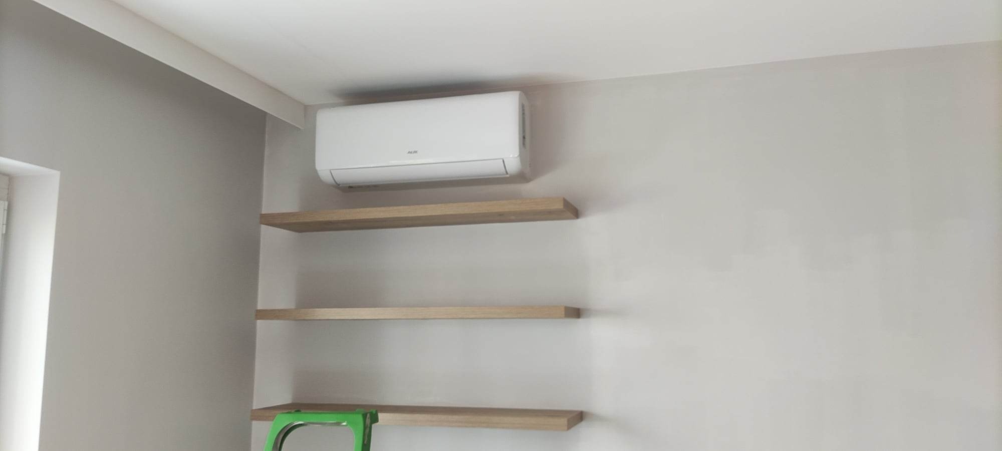Biały klimatyzator AUX zamontowany na ścianie w pokoju, poniżej trzy drewniane półki, zielony fragment drabiny w dolnym rogu.