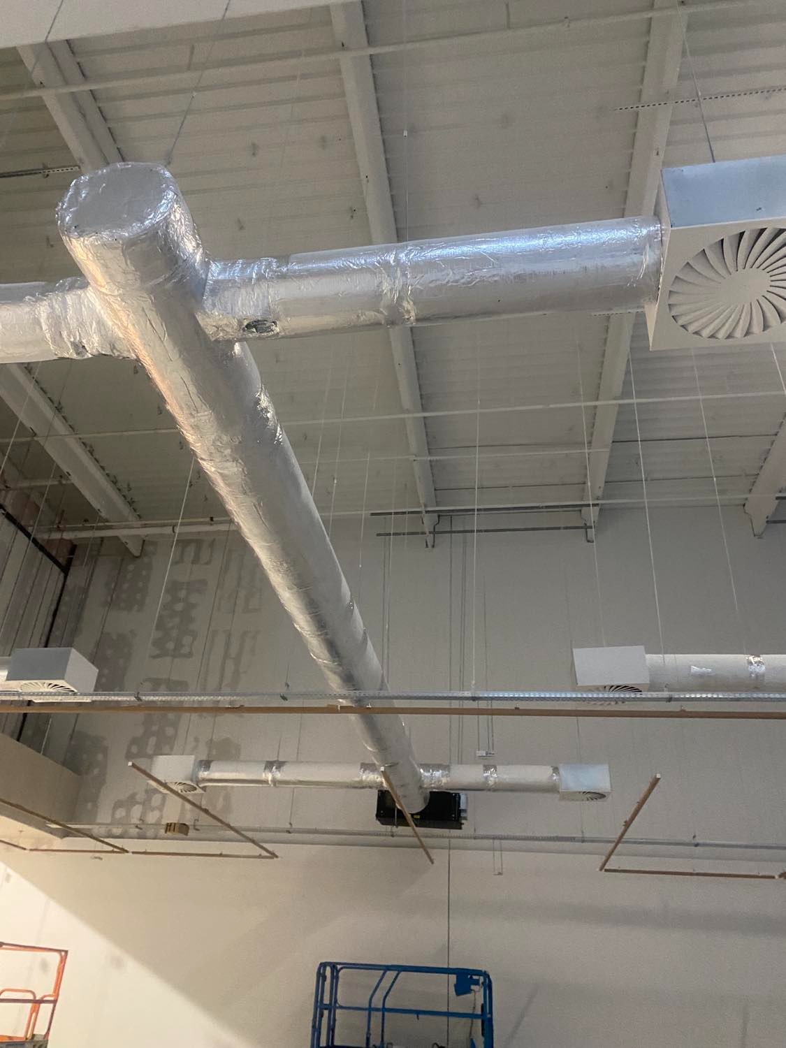 Widok z dołu na system kanałów wentylacyjnych z izolacją aluminiową, kratki wentylacyjne i częściowo widoczne podnośniki koszowe w jasnym pomieszczeniu przemysłowym.